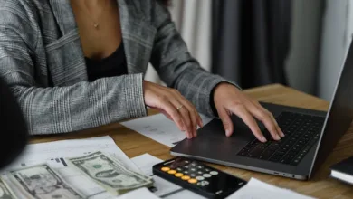 A woman sitting at a desk with a stack of cash next to a laptop and calculating a budget for her move.